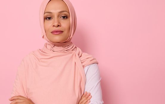 Headshot, close-up face portrait of a gorgeous Arabic Muslim woman in pink hijab posing looking to camera with attractive gaze, confident look and crossed arms, on pink background with copy space