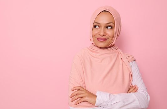 Close-up portrait of a middle aged Arab Muslim gorgeous pretty woman with covered head in hijab looking mysteriously at the side on a pink background with copy space and smiling thoughtfully