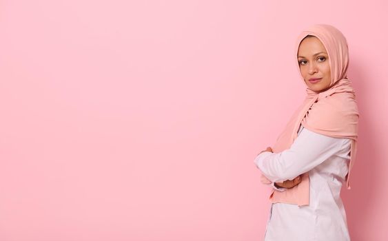 Confident portrait of Arabic muslim beautiful woman with attractive look and gaze, wearing pink hijab and standing sideways against colored background with copy space