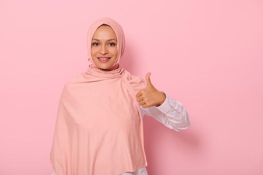 Isolated portrait on pink background with copy space of an adorable and charming Arabic woman in hijab showing a thumb up and smiling with beautiful toothy smile looking at the camera