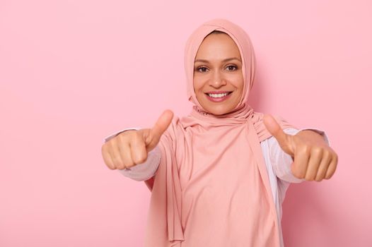 Arab Muslim friendly and charming woman in pink hijab shows thumbs up to the camera, smiles with toothy smile, isolated over colored background with space for text