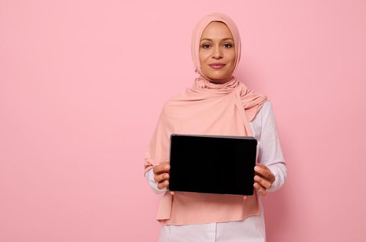 Confident portrait of Arab Muslim beautiful successful woman wearing pink hijab standing against pink background with copy space and showing the empty blank screen of a digital tablet to the camera.