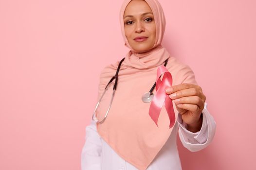 Soft focus on Breast Cancer Awareness Ribbon, in hands of blurred confident Muslim woman doctor in pink hijab, isolated on pink background, copy space. 1 st October, World Day of fight breast cancer