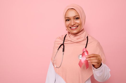 Gorgeous Arab Muslim female doctor in hijab with beautiful toothy smile showing Pink satin Ribbon, isolated on colored background, copy space. World Day of fight Breast Cancer, 1 st October concept