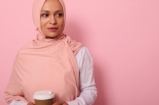Close-up of Arab Muslim beautiful woman wearing pink hijab and white shirt holding a recyclable disposable ecological paper mug in her hands, looking the side posing on colored background, copy space