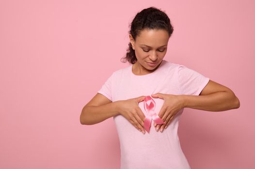 Isolated portrait on colored background of mixed race woman in pink t-shirt, putting her hands on her chest in shape of heart with a pink satin ribbon in the center. World Cancer Awareness Day.