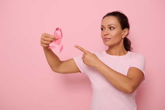 Beautiful mixed race woman wearing pink T-shirt pointing to a pink satin ribbon in her hand, colored background copy space. Concept of 1 st October, International Day of fight Breast Cancer illness