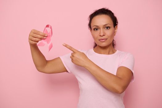 Portrait of serene Middle Eastern ethnicity woman in pink T-shirt pointing to a satin ribbon in her hand, looking at camera, isolated on colored background with copy space. Breast Cancer Day concept