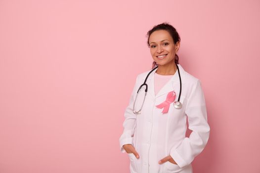 Portrait of African American womandoctor in medical coat and pink ribbon, stethoscope around neck, looking at camera, isolated on colored background, copy space. World Breast Cancer Awareness Day