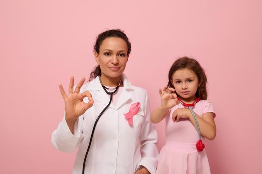 Pretty woman doctor in medical gown and a cute little girl, both with a phonendoscope around their neck, gesticulate with an OK sign to support breast cancer patients and survivors. Medical concept