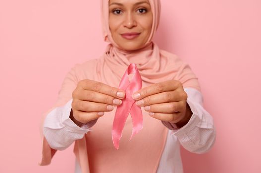 Blurred smiling Muslim woman with covered head in pink hijab holding satin ribbon in her hands. Breast and abdominal cancer awareness, October Pink day, copy space. Breast cancer support concept