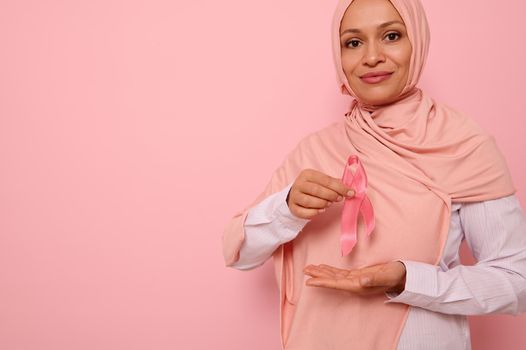 Confident portrait of a mature Arab Muslim woman wearing a hijab holding a pink satin ribbon at chest level to show her support for cancer patients and survivors. World Breast Cancer Awareness Day.