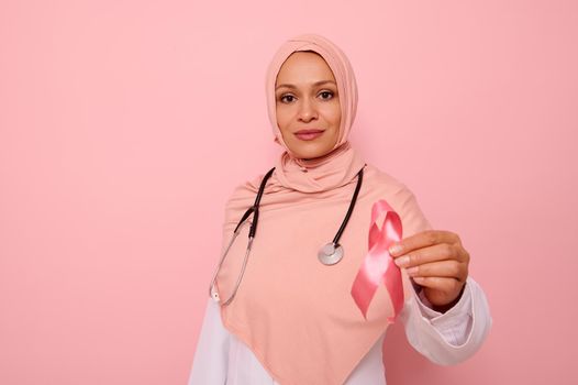 Gorgeous serene Arab Muslim female doctor in pink hijab holding Pink satin Ribbon, looking at camera, isolated on colored background, copy space. World Day of fight Breast Cancer, 1 st October concept