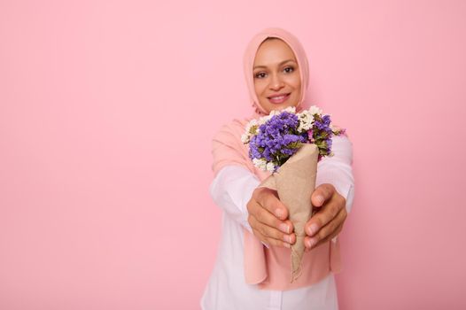 Focus on a beautiful simple bouquet of meadow wildflowers in purple tones, wrapped in brown craft paper in the hands of an attractive smiling Muslim woman wearing a hijab. Pink background copy space