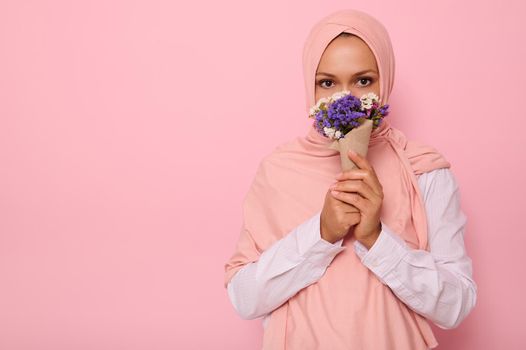 Confident portrait of young charming Arab Muslim woman in pink hijab with beautiful dark eyes, attractive gaze, looking at camera, covers half of her face and mouth with a craft bouquet of wildflowers