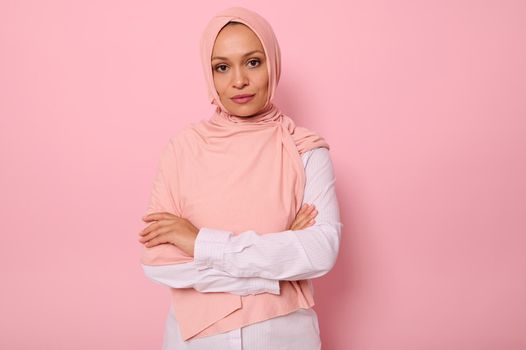 Waist-length portrait of beautiful Arabic Muslim woman in pink hijab posing looking to camera with attractive gaze, confident look and crossed arms, on pink background with copy space