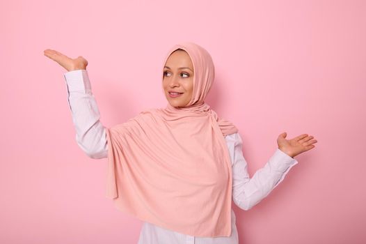 Beautiful young Arab Muslim woman raising hand palm up while looking at it with smile. Isolated background with copy space