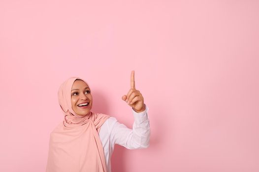 Muslim woman of Middle Eastern ethnicity dressed in religious outfit and covered head with hijab ethnicity smiles with toothy smile and looks up pointing her index finger on pink background copy space