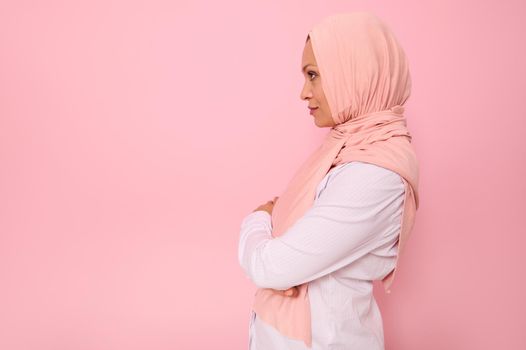 Confident close-up portrait of Arab muslim confident woman with attractive gaze , wearing a pink hijab looking confidently in front of herself standing sideways on colored background with copy space
