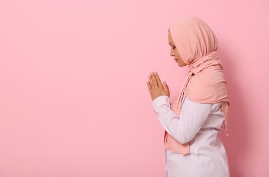 Side portrait of a serene Muslim Arab woman in pink hijab and strict outfit praying, performing namaz, isolated on colored background with space for text
