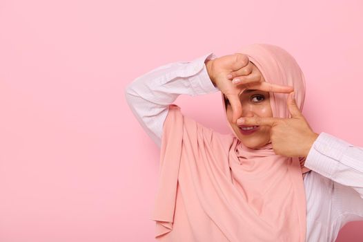 Headshot, close-up face portrait of a young Muslim pretty woman of Arabic ethnicity, wearing hijab and looking at the camera through a finger frame, isolated on colored background with copy space