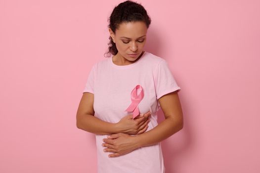 Mixed race woman puts hands under pink ribbon on her pink Shirt, for breast cancer campaign, supporting Breast Cancer Awareness. Concept of 1 st October Pink Month and women's health care. Copy space