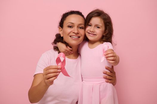 Mixed race young woman hugs her daughter, holding pink ribbon, cute smiles looking at camera, isolated on colored background with copy space. International Day of fight against a Breast Cancer disease