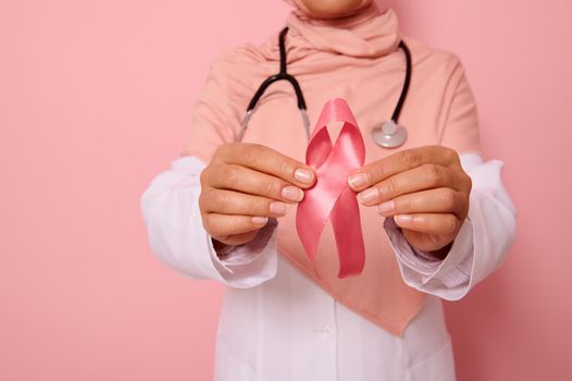 Focus on hand of doctor in pink hijab, holding pink ribbon. World Cancer Day. Breast and abdominal cancer awareness, October Pink day, colored background, copy space. Breast cancer support concept.