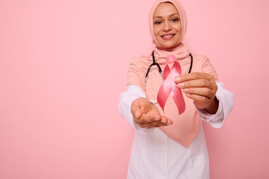 Focus on Pink Breast Cancer Awareness ribbon. Muslim doctor in pink hijab posing on pink background, showing support, solidarity to patients cancer survivors. World Day of fight cancer Medical concept