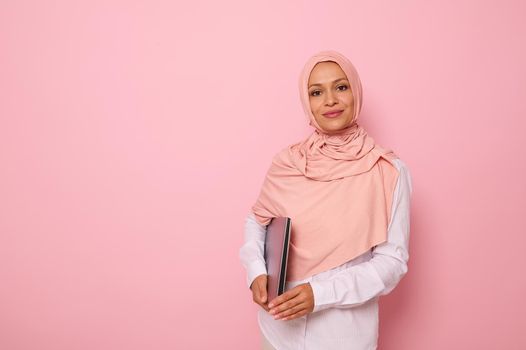 Isolated confident business portrait on colored background of Arab muslim pretty woman with covered head in pink hijab, wearing white shirt, looking at camera with a laptop in hands. Space for text