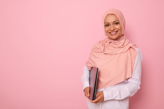 Gorgeous Muslim woman in stylish traditional religious outfit with covered head in pink hijab smiling with toothy smile posing against colored background with a laptop in hands. Copy space