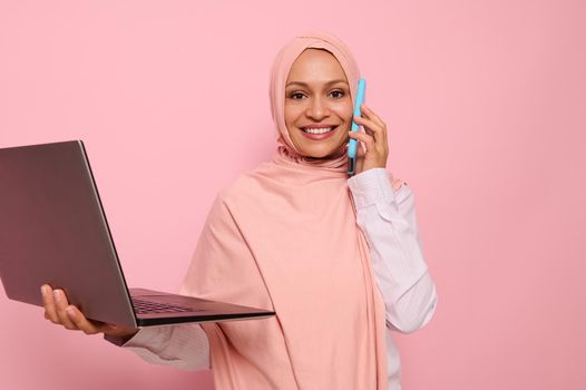 Portrait of a beautiful Arab Muslim woman in pink hijab talks on mobile phone, looks at camera, smiles with toothy smile and holds a laptop. Business concept on pink pastel background with copy space.