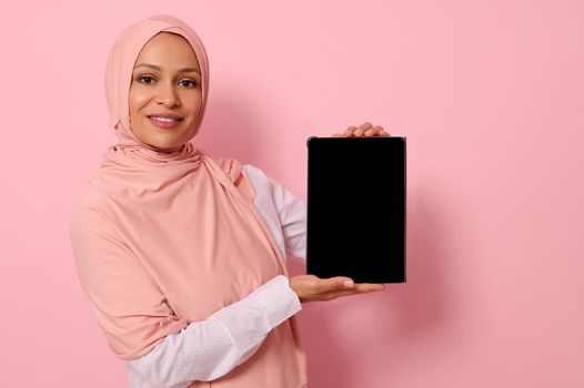 Confident Muslim woman of Arab or Middle Eastern ethnicity in hijab holding vertically a digital tablet and showing its blank screen, smiles looking at camera, isolated on pink background, copy space