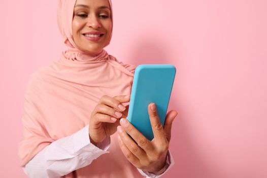 Close-up portrait of confident Arab Muslim mature pretty woman in strict religious outfit and covered head in pink hijab texting message on a mobile phone in her hands, colored background, copy space