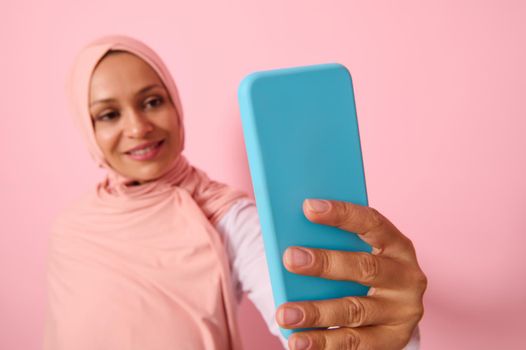 Soft focus on the smartphone in blue cover in outstretched arms of Arab Muslim woman wearing traditional religious islamic outfit, pink hijab,and smiles with toothy smile making a selfie, self-portrait