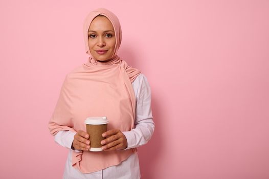 Arab Muslim beautiful woman wearing pink hijab and white shirt holding a recyclable disposable ecological paper mug in her hands, looking at camera posing on colored background with copy space