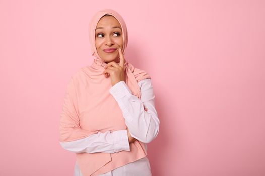 Adorable young Muslim beautiful gorgeous woman with a covered head in hijab looks mysteriously at a pink background with copy space and puts her finger to the corner of her lips, smiling thoughtfully