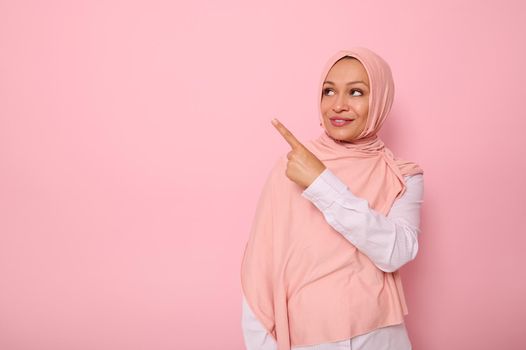 Gorgeous Arab Muslim woman with covered head in hijab looking at the pink background and pointing her index finger indicating the copy space for text. Business concepts with a religious beautiful lady