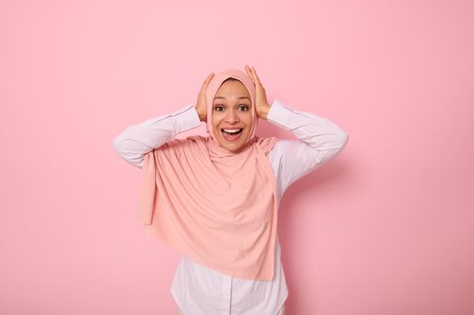 Happy religious Muslim woman in pink hijab holding her head with hands and smiling with toothy smile looking at camera with surprised look, isolated on colored background with copy space.