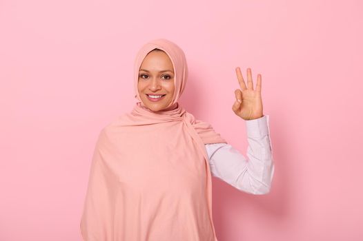 Charming joyful young Arab Muslim pretty woman dressed traditional religious attire and covered head in hijab showing OK sign, looking at the camera, standing against pink background with copy space
