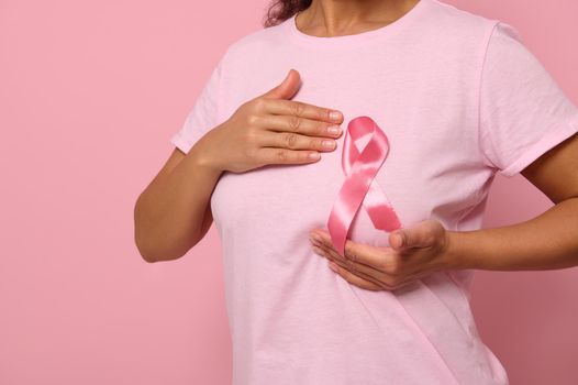 Close-up woman puts hands under pink ribbon on her T Shirt, supporting Breast Cancer Awareness campaign. Concept of 1 st October Pink Month, women's health care, colorecd background, copy space