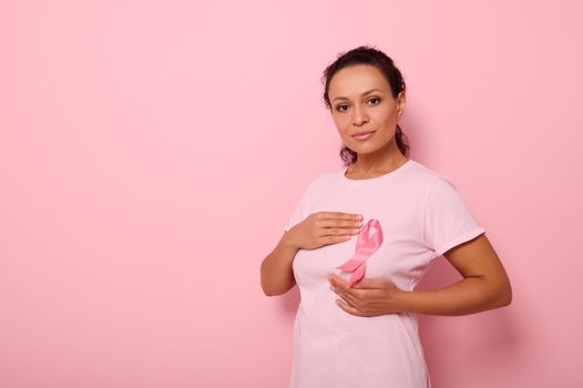 Mixed race woman puts hands around pink ribbon on her pink T Shirt, for breast cancer campaign, supporting Breast Cancer Awareness. Concept of 1 st October Pink Month and women's health care