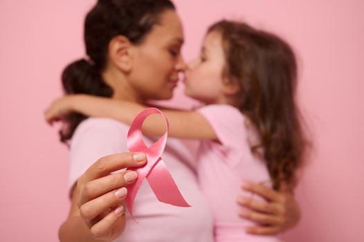 Soft focus on a pink satin ribbon, symbol of International Breast Cancer Awareness Day, against blurred background of loving mother and lovely daughter. Female healthcare and medical education concept