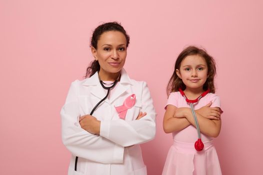 Gorgeous woman doctor in white medical gown stands next to a cute little girl in pink, both with crosses arms on chest and wearing a pink ribbon, symbol of Breast Cancer Awareness Day. Medical concept