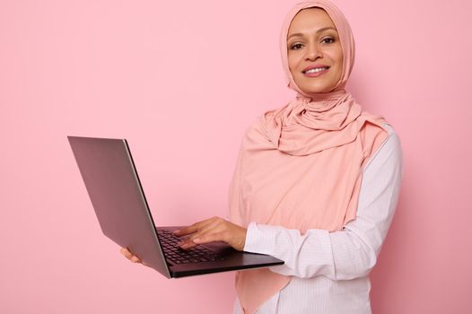 Business portrait of successful programmer Muslim woman in pink hijab standing against a colored background with an open silver laptop in hands and typing text, smiling looking at camera, copy space.
