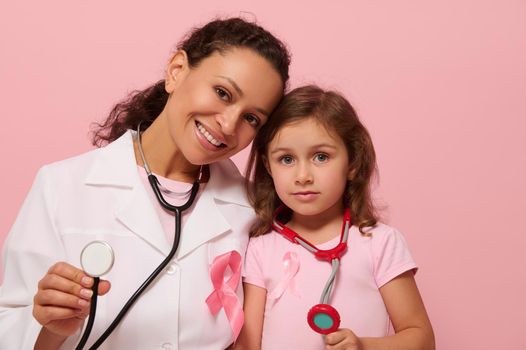 Smiling female doctor and cute little girl, both with Breast Cancer Awareness pink ribbon and phonendoscope around their neck, showing stethoscope to camera, isolated on pink background, copy space.
