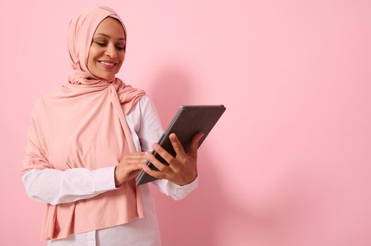 Beautiful smiling Arab Muslim woman with covered head in pink hijab headscarf, posing against a colored background with a digital tablet in hands, copy space for text. Advertising isolated portrait