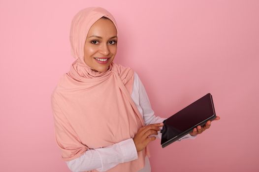 Arab Muslim woman in pink hijab smiles with toothy smile looking at camera, posing on pastel colored background with digital tablet in hands. Isolated portrait for advertising with copy space for text