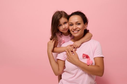 Woman's health , medical concept. Serene woman and girl in pink attire with Breast Cancer Awareness ribbon, daughter hugging her mom, holding by hands, looking at camera, supporting cancer patients.