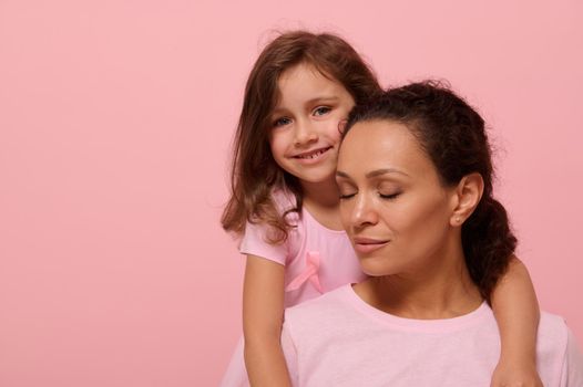 Beautiful baby girl in pink dress with pink ribbon tenderly hugs her mother with closed eyes, looking at camera, showing support to cancer patients and survivors, pink background with copy space